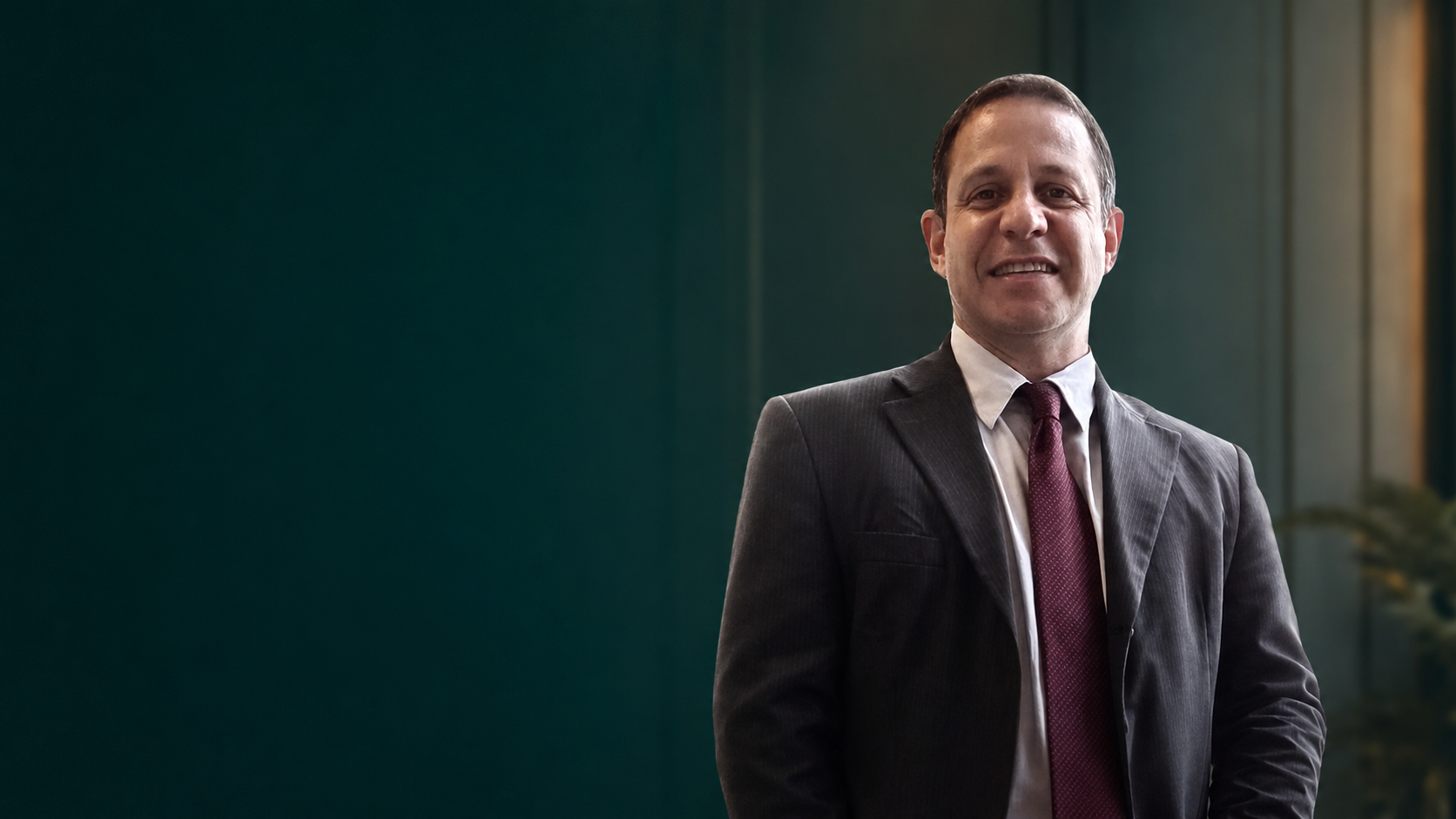 Retrato de advogado trabalhista sênior sorrindo, terno cinza escuro e gravata bordô, em pé diante de parede verde-petróleo, iluminação suave e profissional, fotografia corporativa realista